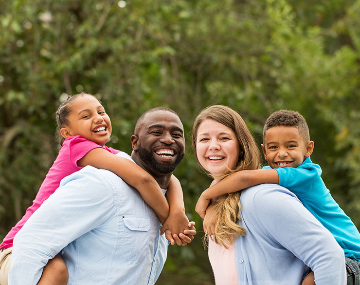 Family of four smiling at the camera, they are outdoors in a park